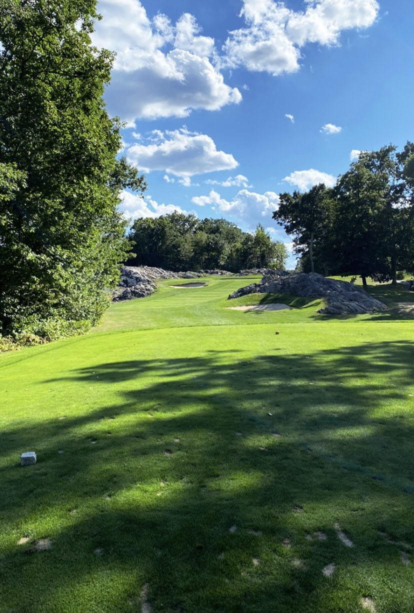 Tee view at Breakfast Hill Golf Club in Greenland, New Hampshire, looking up a shaded fairway toward a bunkered green flanked by granite rock outcroppings and hardwoods