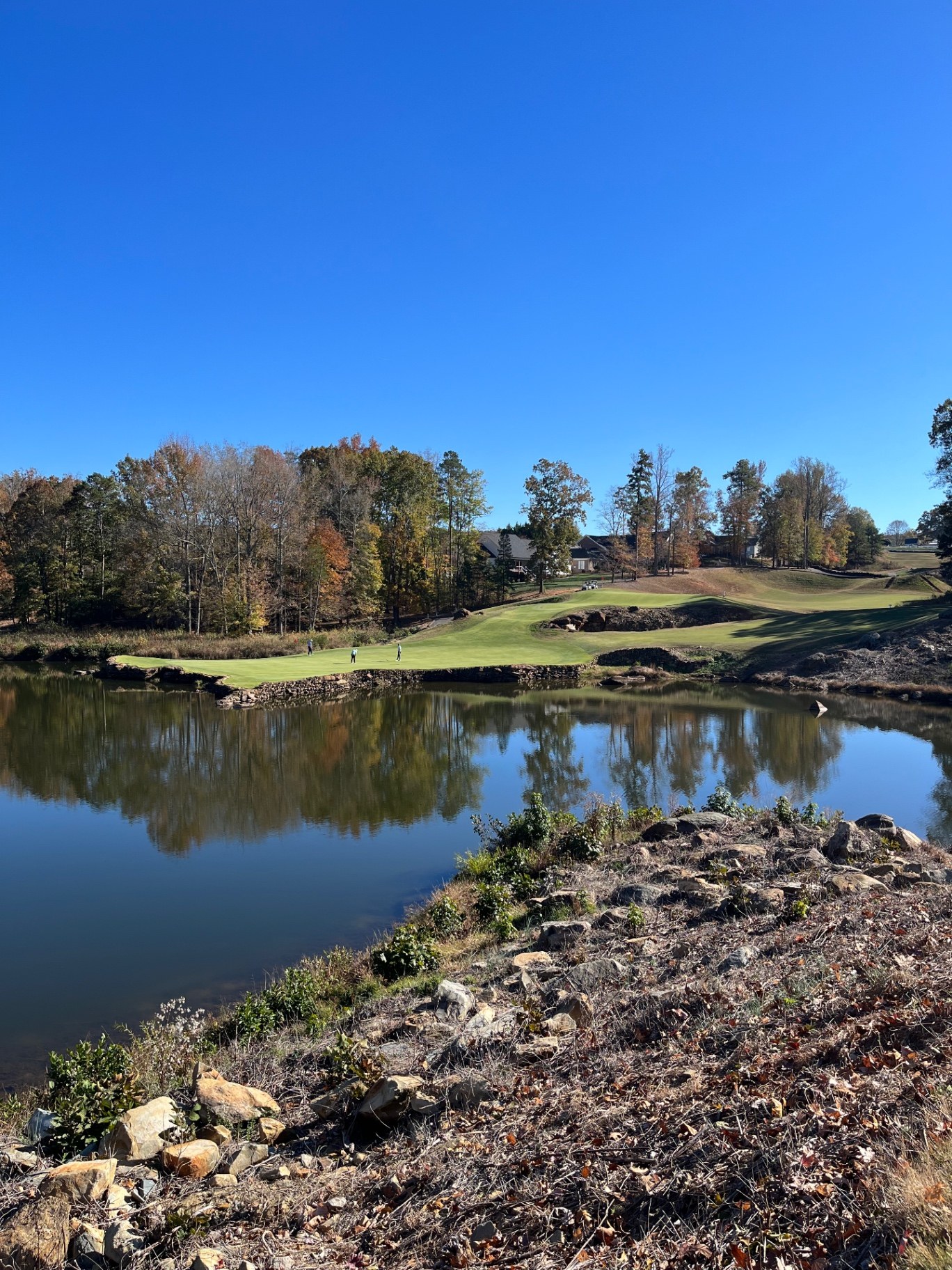 Green across a pond at Tot Hill Farm Golf Club in Asheboro, North Carolina, framed by rocky terrain and fall foliage under a deep blue sky