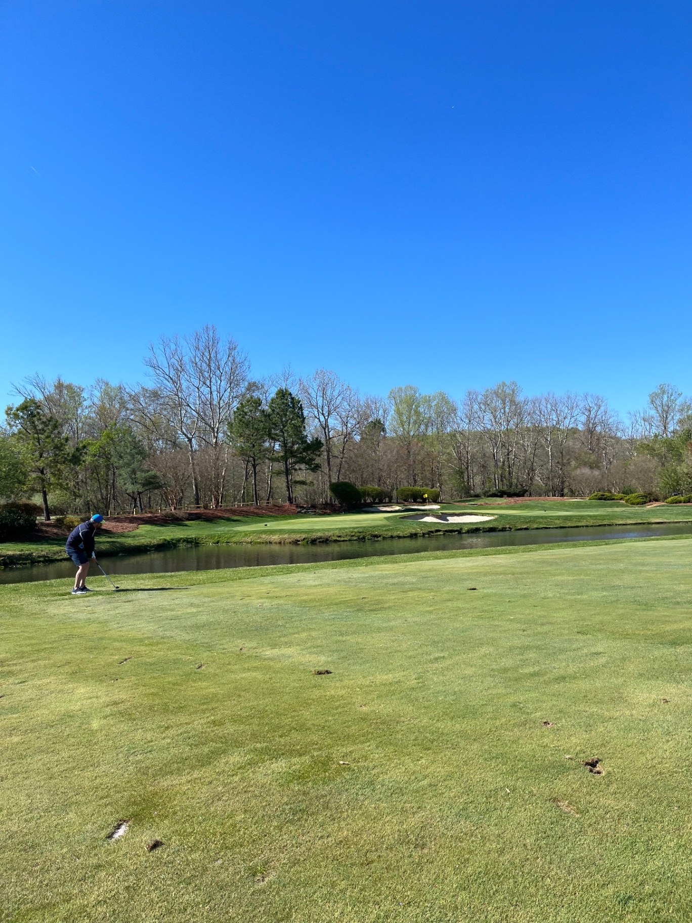 Golfer playing an approach shot across a creek to a bunkered green at Renditions Golf Course in Davidsonville, Maryland under a clear blue sky