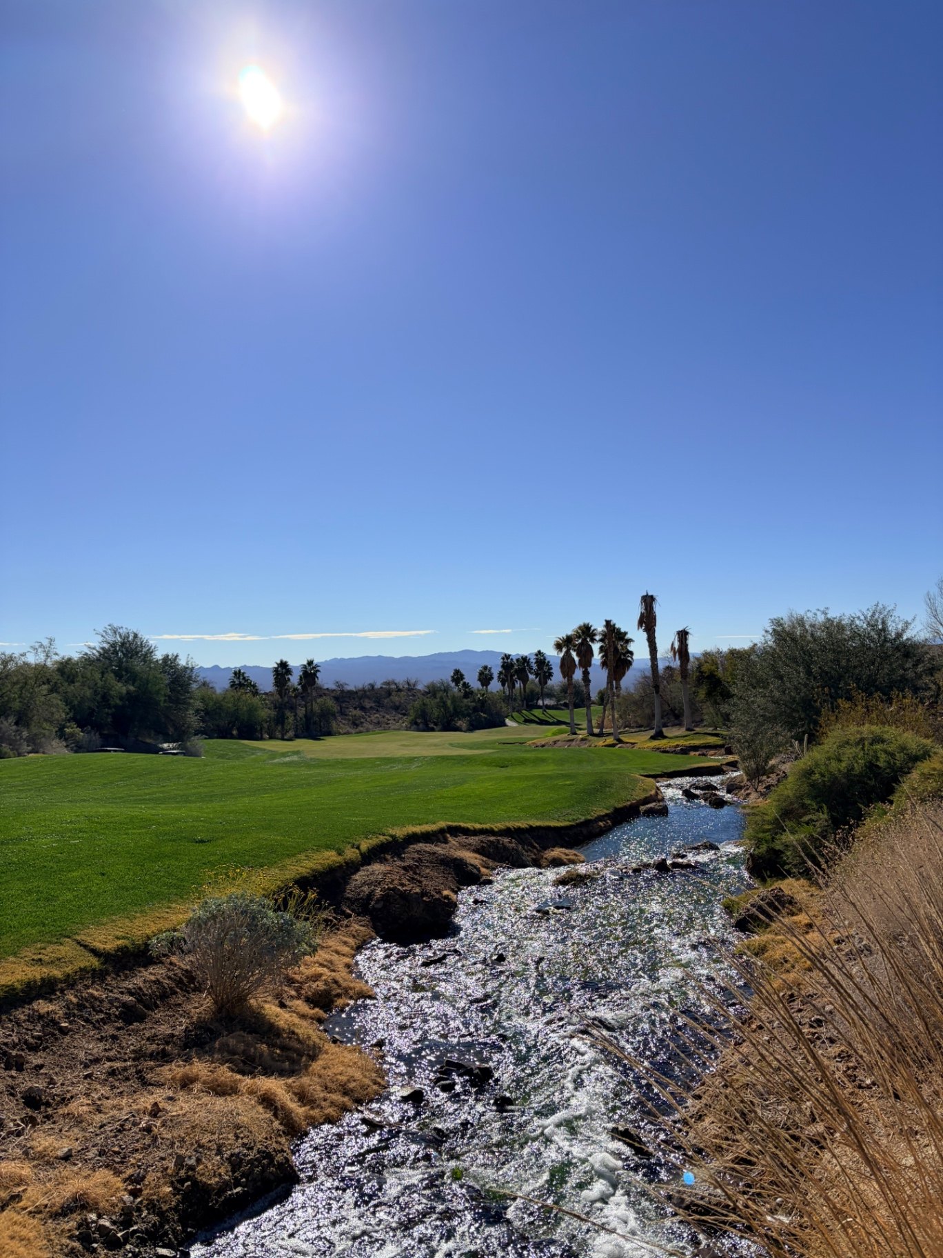 Desert fairway at Cascata beside a rushing rock-lined stream with palms and distant mountains