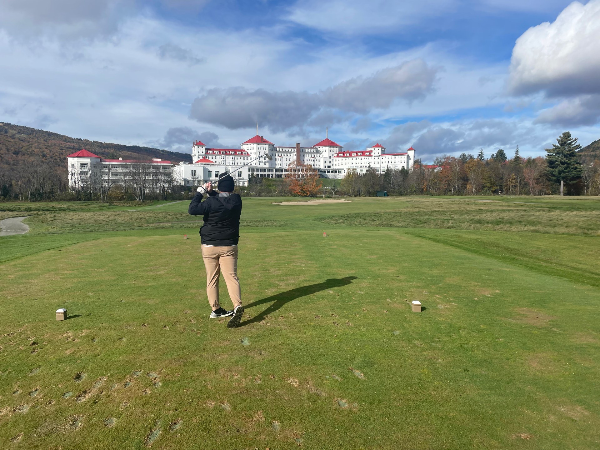 Golfer finishing a tee shot on the Mount Washington Course at Omni Mount Washington Resort, with the white red-roofed hotel and White Mountains beyond