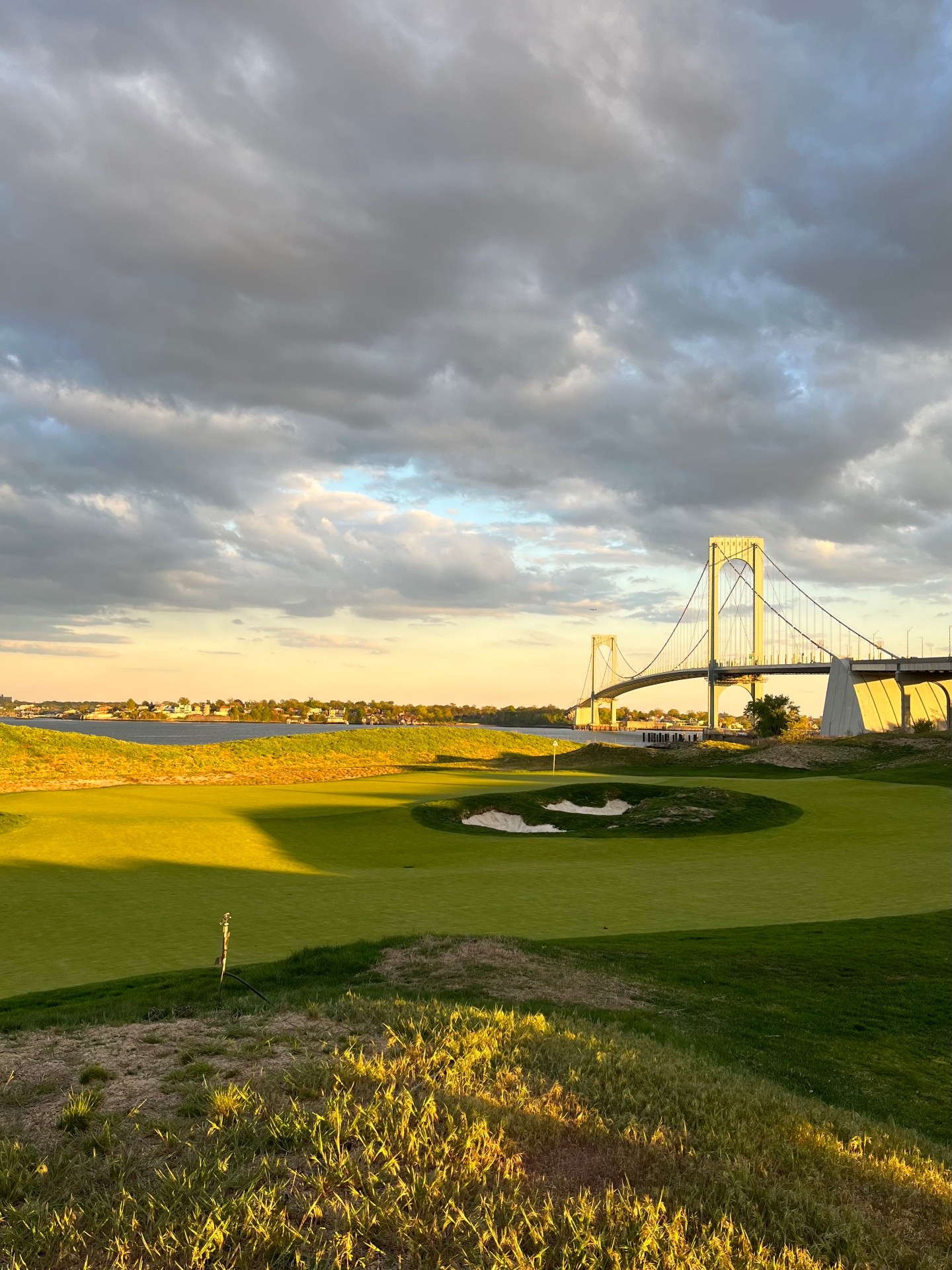 Green with a deep bunker at Bally's Golf Links at Ferry Point, with the Bronx-Whitestone Bridge and East River in golden evening light