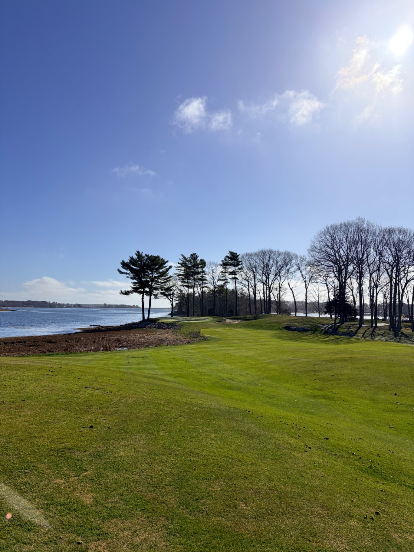 Coastal fairway at Wentworth by the Sea Country Club bending along tidal water with pines and bare trees under blue sky