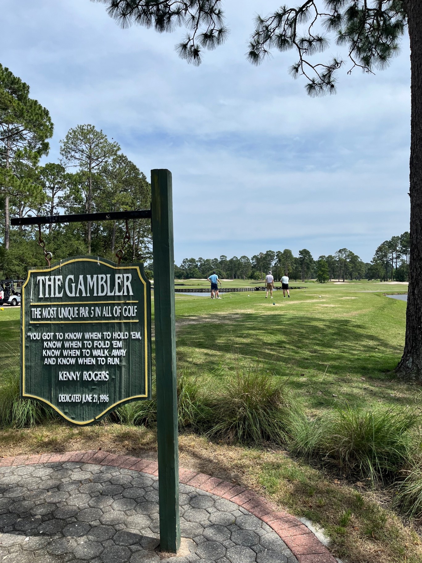 The Gambler tee sign at Myrtle Beach National King's North, with golfers on the par-5 tee beyond it and pines lining the fairway