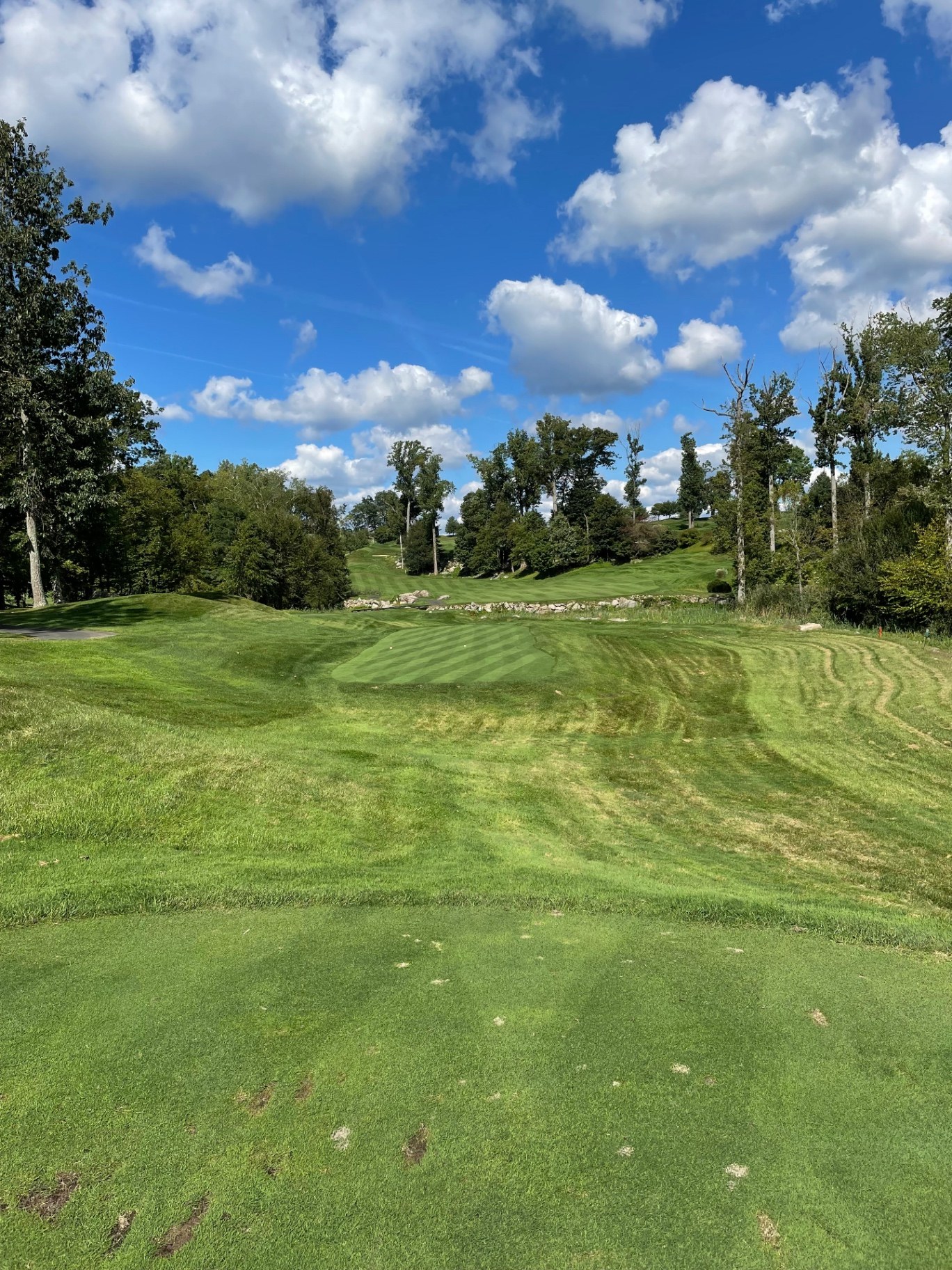 View from an elevated tee across rugged rock outcrops and rolling contours toward a striped green surrounded by hardwoods at Pound Ridge Golf Club