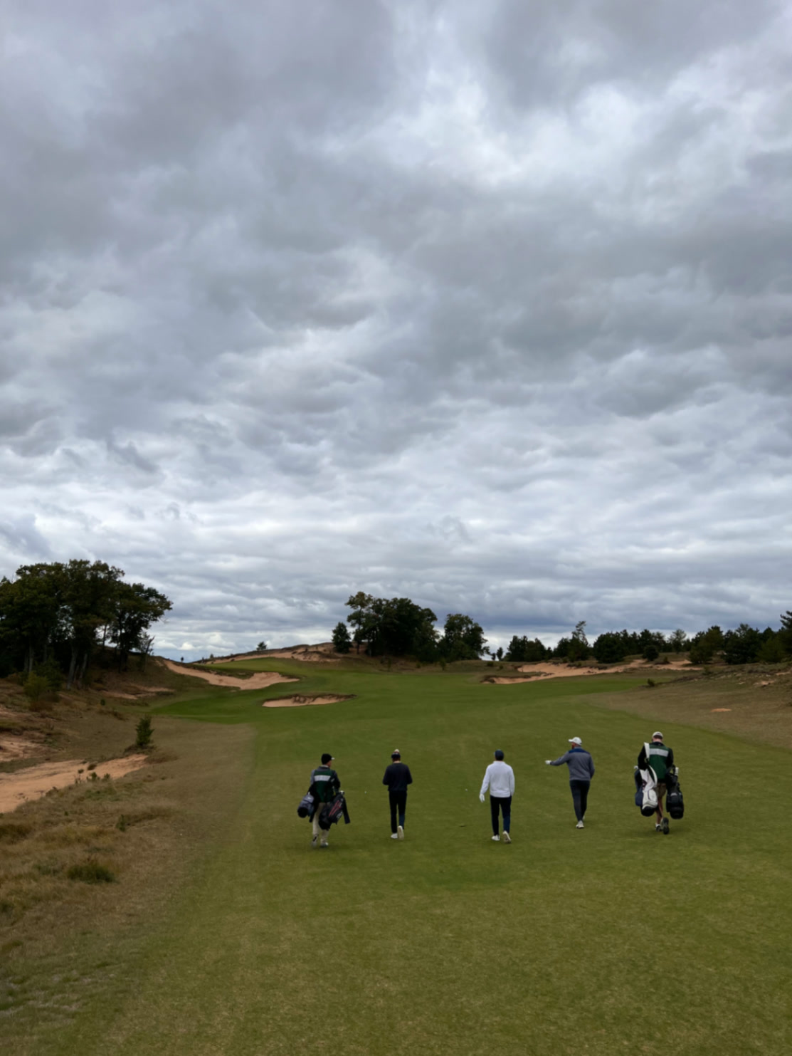 A foursome walking a wide sand-framed fairway under heavy grey clouds at Sand Valley Golf Resort in Nekoosa, Wisconsin